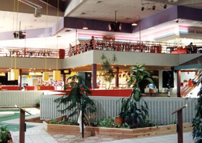 Interior of a multi-level shopping mall with sparse foot traffic, featuring indoor plants, seating areas, and retail stores in the background.