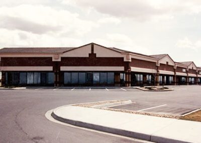 A suburban strip mall with multiple vacant storefronts under a cloudy sky, seen from a curved road in the foreground.