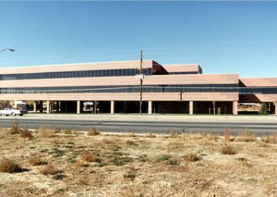 A large, multi-story brown office building beside a road with sparse traffic and an empty grassy field in the foreground.