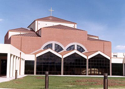 Modern church building with large glass windows and multiple arches, topped by a cross under a blue sky.