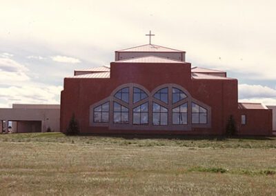 A modern church with a large arched window and a cross on top, set against a partly cloudy sky.