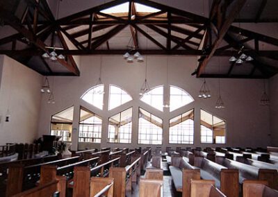 Interior of a church with wooden pews, large arched windows, and exposed beam ceiling, illuminated by hanging lights.