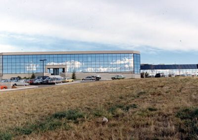 Modern office building with reflective glass facade, surrounded by a parked cars and a grassy foreground under a cloudy sky.