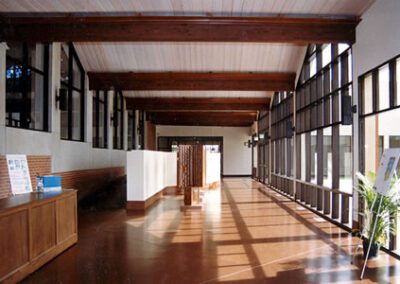 Empty school hallway with wooden floors and ceiling, lined by open black framed windows and white walls, illuminated by natural light.